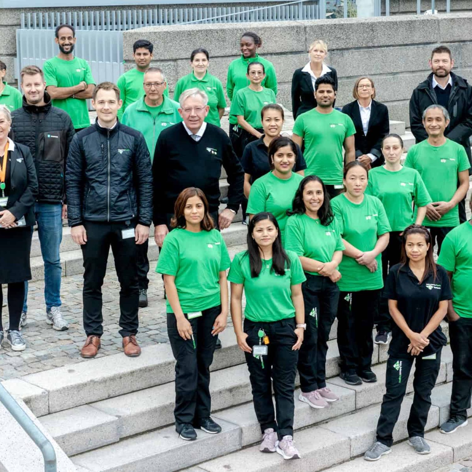 Teamfoto af rengøringspersonale i grønne uniformer foran en trappe.